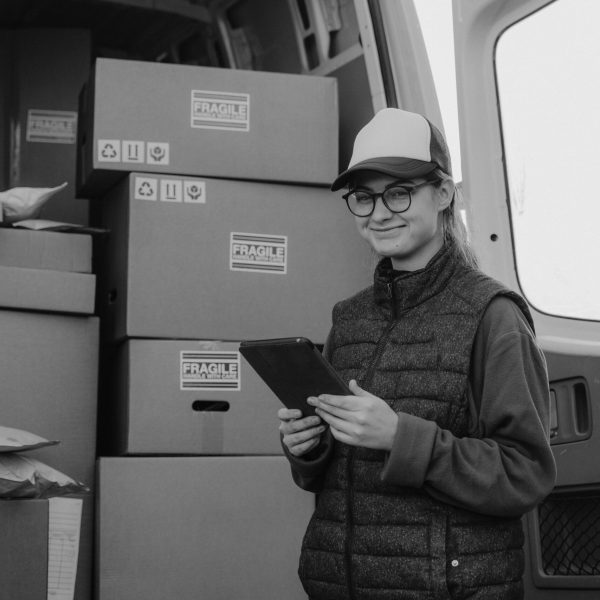 A delivery woman holding a tablet stands next to van full of parcels.